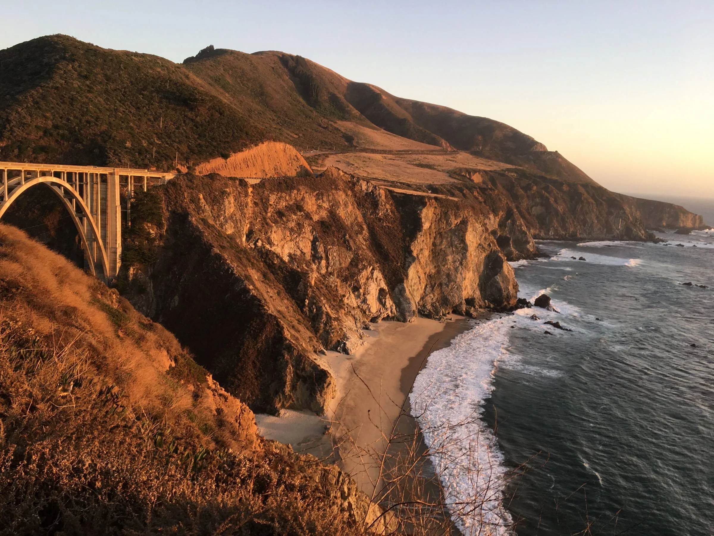 An arched concrete bridge on the cliffs above a Pacific cove in low golden light