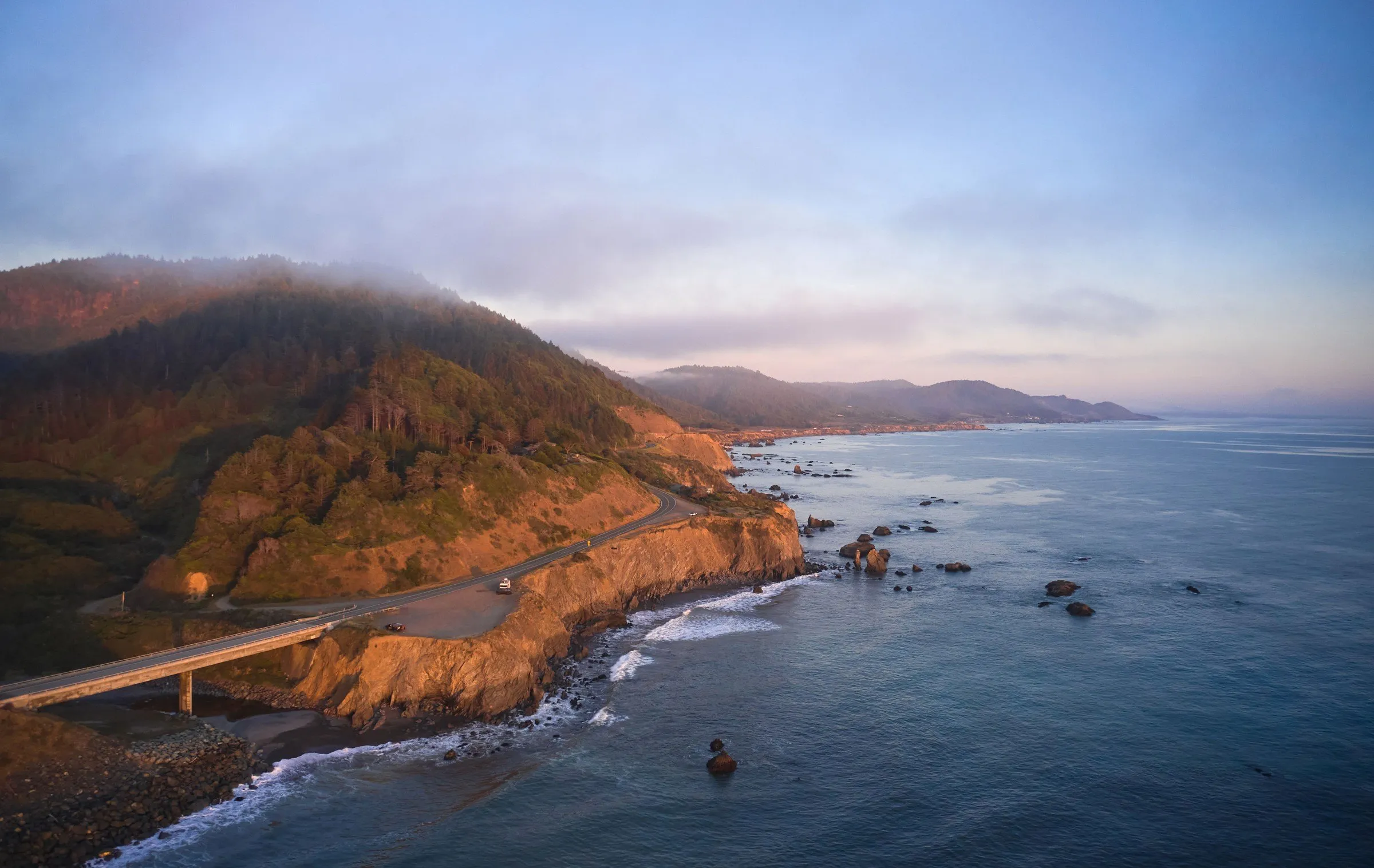 A coast road running along golden cliffs above the Pacific in early evening light