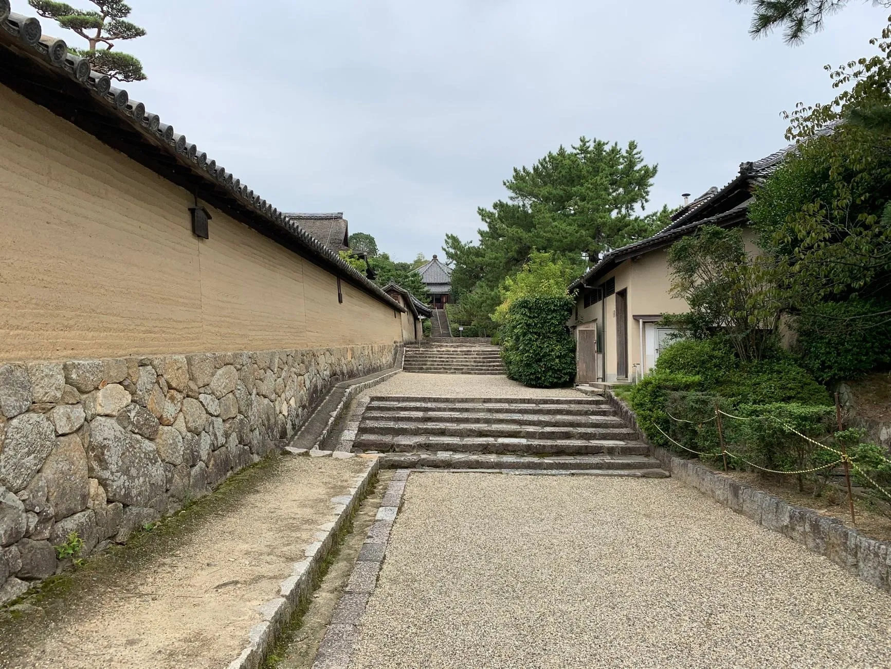A long earthen temple wall beside a gravel path and stone steps under pine trees